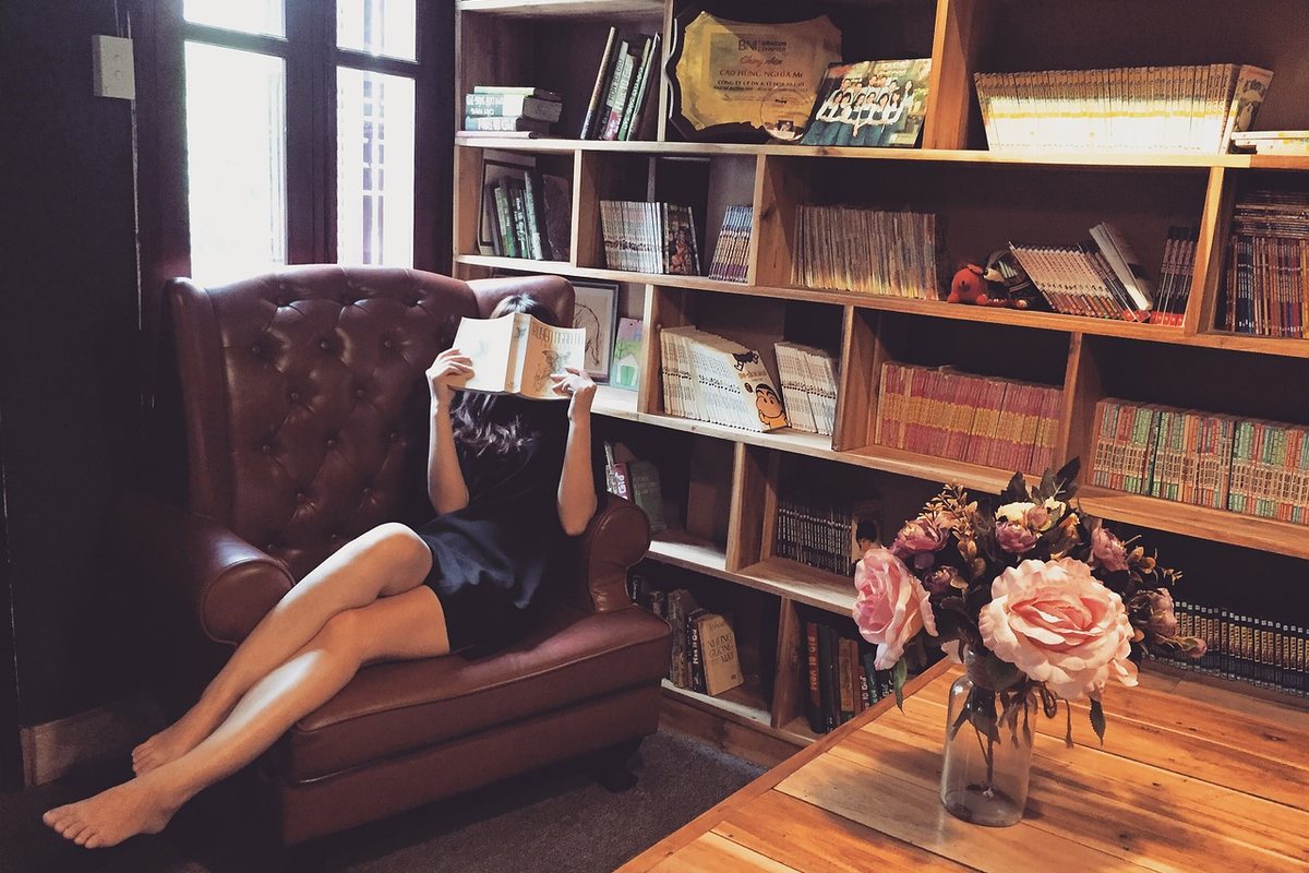 A person reading in a brown buttoned leather armchair in front of full bookshelves.
