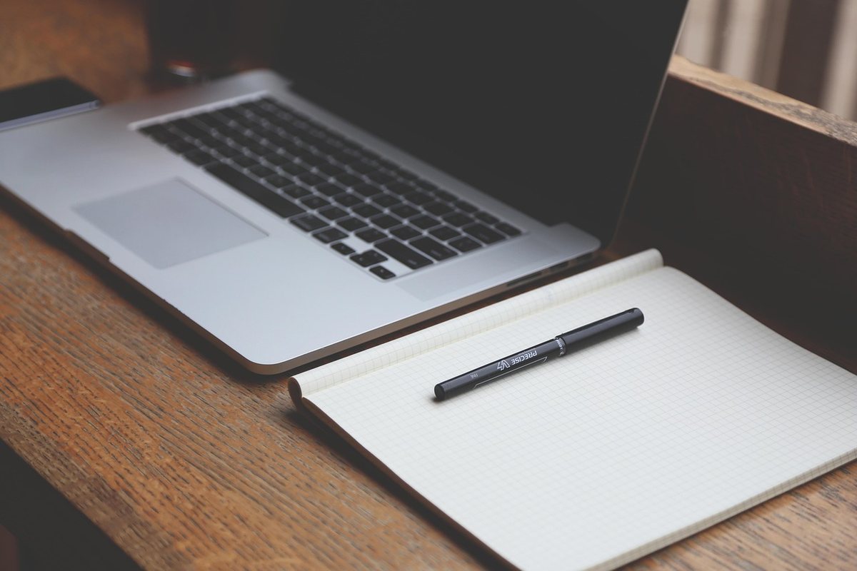 A laptop and an open notebook on a plain wooden table top.