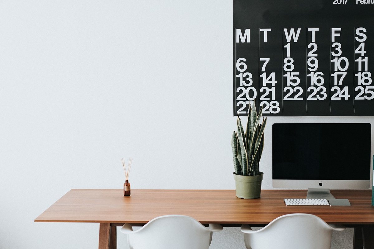 A wooden desk against a wall with a monitor, a plant and a wall calendar above it.