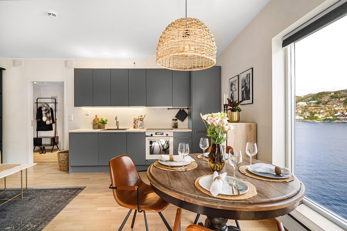 A round wooden dining table with leather-seat chairs in a grey kitchen beside a window.