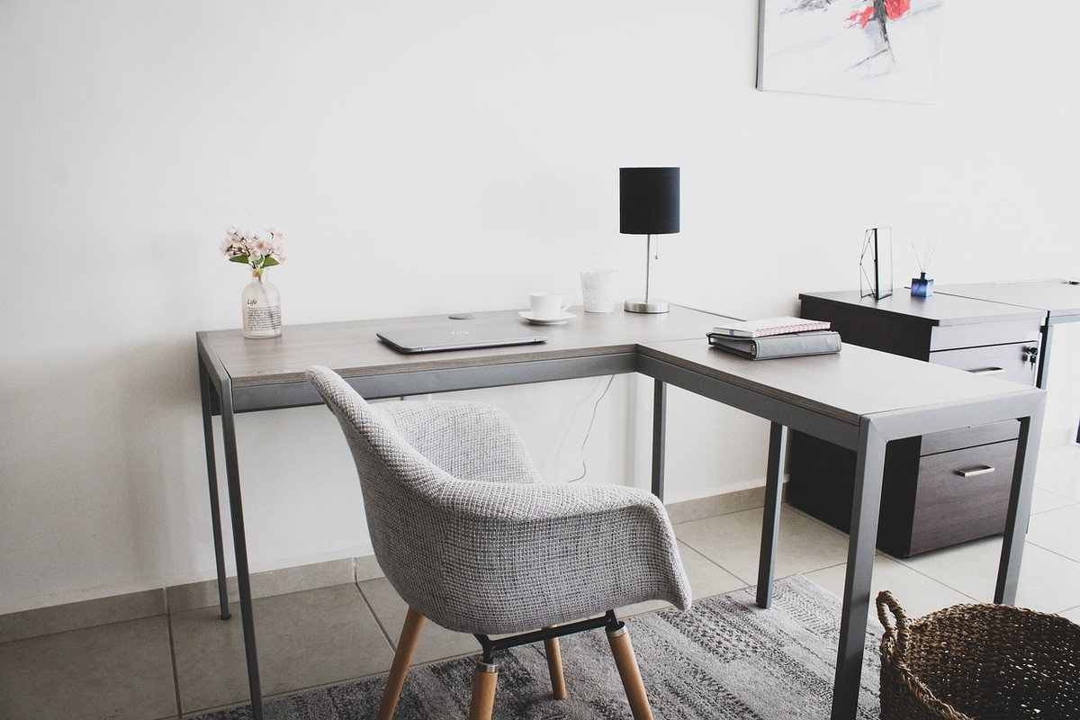 A home office with an L-shaped wooden desk, a task chair and a window behind it.